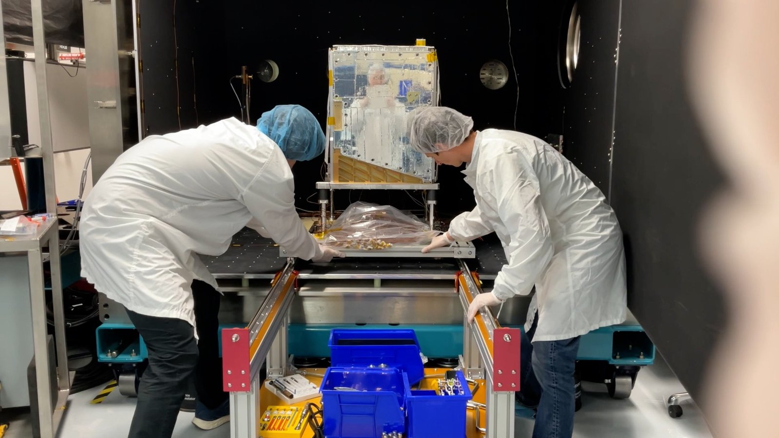 Engineers in cleanroom suits loading the satellite into a thermal vacuum chamber